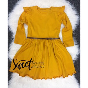 May include: A mustard yellow long-sleeved dress with a ruffled neckline and hem. The dress has a brown belt and the text "Sweet White Peony" is printed on the front.