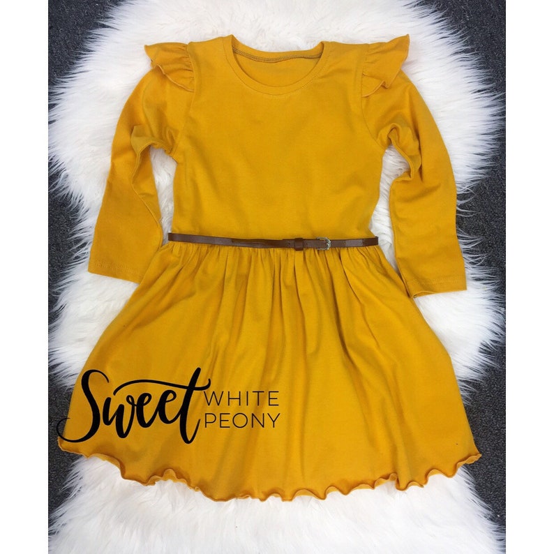 May include: A mustard yellow long-sleeved dress with a ruffled neckline and hem. The dress has a brown belt and the text "Sweet White Peony" is printed on the front.