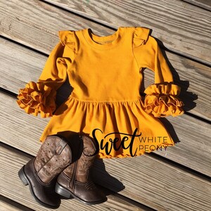 May include: A mustard yellow long-sleeved dress with ruffled sleeves and a ruffled hem. The dress is on a wooden surface with a pair of brown cowboy boots.