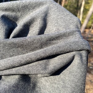 May include: Close-up of a folded, dark gray fabric. The material appears to be soft and slightly textured, with visible folds and creases. The fabric is a solid, neutral color, suitable for various clothing items.