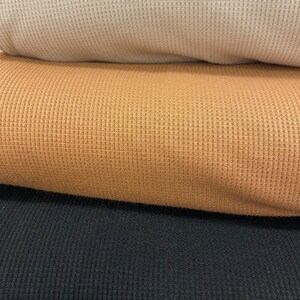 May include: Close-up of three rolled waffle-knit fabrics in neutral colors. The top fabric is a light beige, followed by a golden brown, and the bottom is black. The textured knit creates a grid-like pattern.
