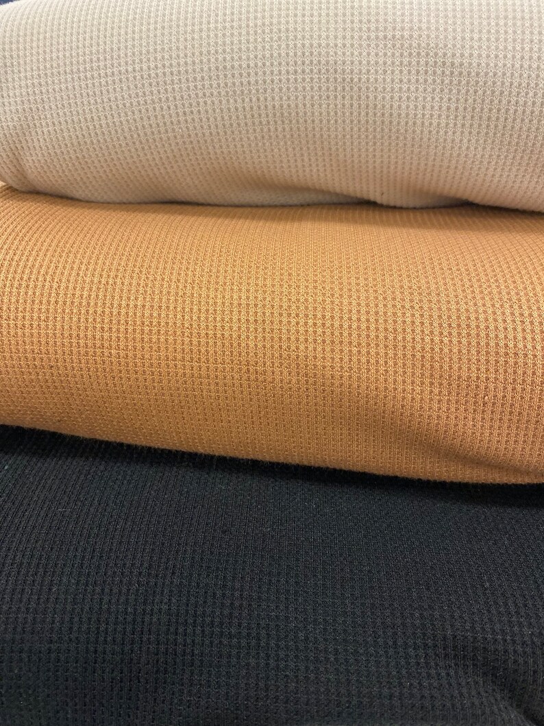 May include: Close-up of three rolled waffle-knit fabrics in neutral colors. The top fabric is a light beige, followed by a golden brown, and the bottom is black. The textured knit creates a grid-like pattern.
