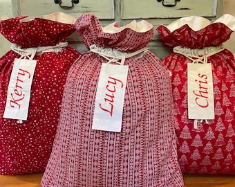 May include: Three red Christmas gift bags with white trim and drawstrings. The bags feature star, line, and Christmas tree patterns. Each bag has a tag with a name: Kerry, Lucy, and Chris.