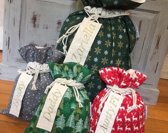 May include: Four festive Christmas gift bags with drawstring tops. The bags display various patterns: a grey bag with white stars, a green bag with trees, a red bag with reindeer, and a green bag with snowflakes. Each bag has a tag with a name embroidered in gold.