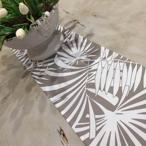 May include: A rectangular table runner featuring a taupe background and a white palm leaf pattern. The runner is displayed on a light-coloured wooden surface. A white vase with white flowers is partially visible in the upper left corner.