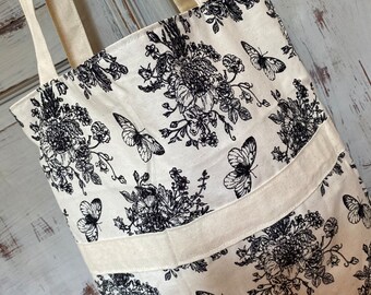 May include: Cream tote bag featuring a black floral and butterfly design. The bag has cream-coloured handles and a cream-coloured band across the middle. The bag is made of a durable fabric.
