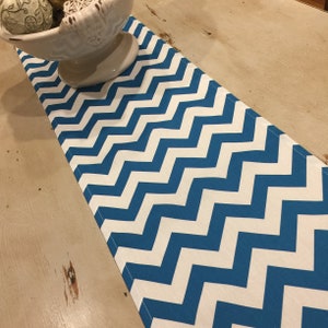 May include: A blue and white chevron patterned table runner. The runner is made of fabric and is placed on a light-colored wooden table. A white bowl with decorative spheres sits on the table.