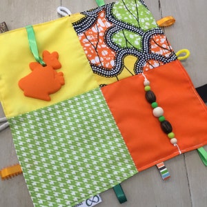 May include: A colorful baby sensory toy with various fabric patterns, including yellow, green houndstooth, and orange. It features an orange animal-shaped teether and a string of wooden beads. Ribbons and loops are attached for tactile exploration.
