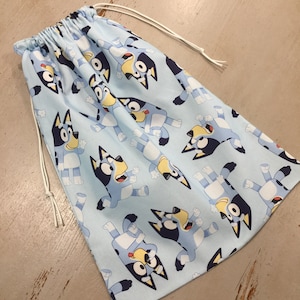 May include: A light blue drawstring bag featuring a repeating pattern of a cartoon dog character in various poses. The bag has a white drawstring closure. The dog character is blue, black, and yellow.