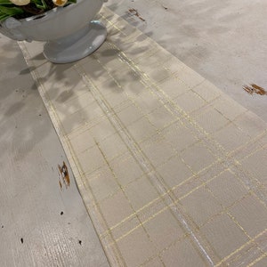 May include: A cream-coloured table runner with a gold metallic plaid pattern. The rectangular runner is made of fabric. Gold lines form a grid design. It's placed on a distressed, light-coloured wooden table. The runner's length is approximately 150 cm.