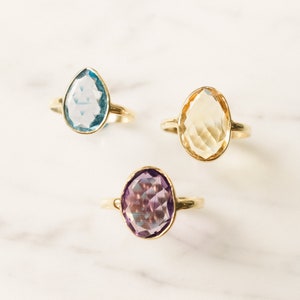 May include: Three gold rings with different colored gemstones. The rings feature a light blue, a yellow, and a purple gemstone.