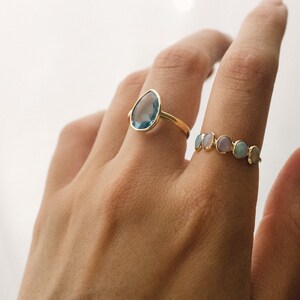 May include: Two gold rings with gemstones. The first ring features a teardrop-shaped blue gemstone. The second ring has a row of small, round, multi-colored gemstones.