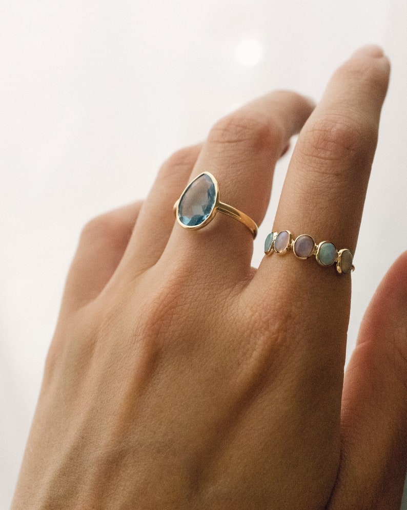 May include: Two gold rings with gemstones. The first ring features a teardrop-shaped blue gemstone. The second ring has a row of small, round, multi-colored gemstones.