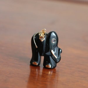 May include: A small, black elephant figurine with gold accents on its feet and a gold loop for hanging. The elephant has a polished finish and a small, red eye. The figurine is likely a pendant or charm.