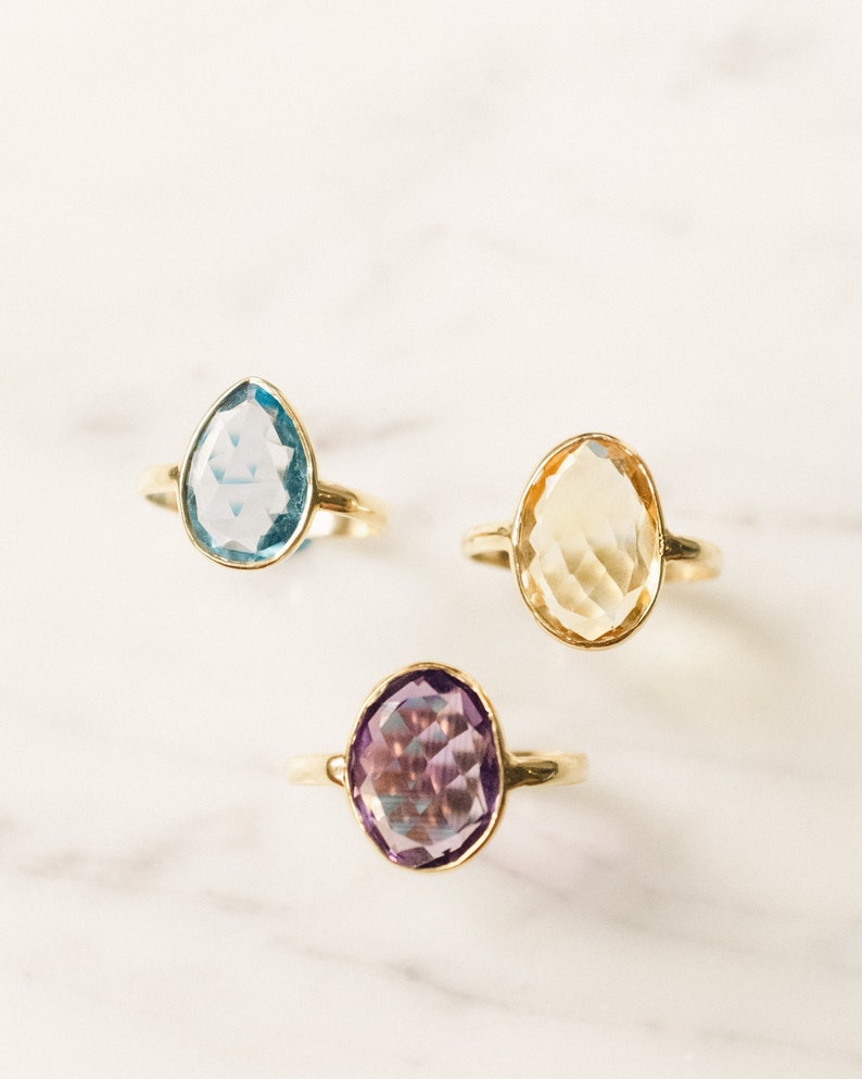 May include: Three gold rings with different colored gemstones. The rings feature a light blue, a yellow, and a purple gemstone.