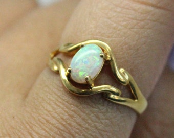 May include: A gold ring featuring an oval opal gemstone. The opal has a milky white base with flashes of green and blue. The ring's band has a flowing, organic design.