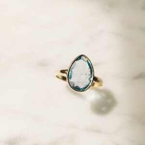 May include: A gold ring with a light blue gemstone. The gemstone is a teardrop shape and has a faceted cut.