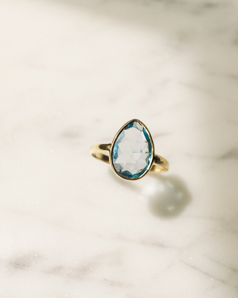 May include: A gold ring with a light blue gemstone. The gemstone is a teardrop shape and has a faceted cut.