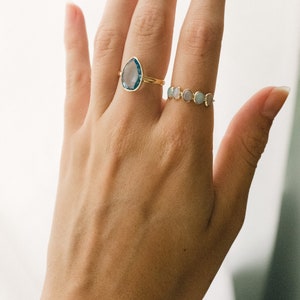 May include: A hand wearing two gold rings. The first ring features a teardrop-shaped blue gemstone. The second ring has a band of small, round, pastel-colored gemstones.