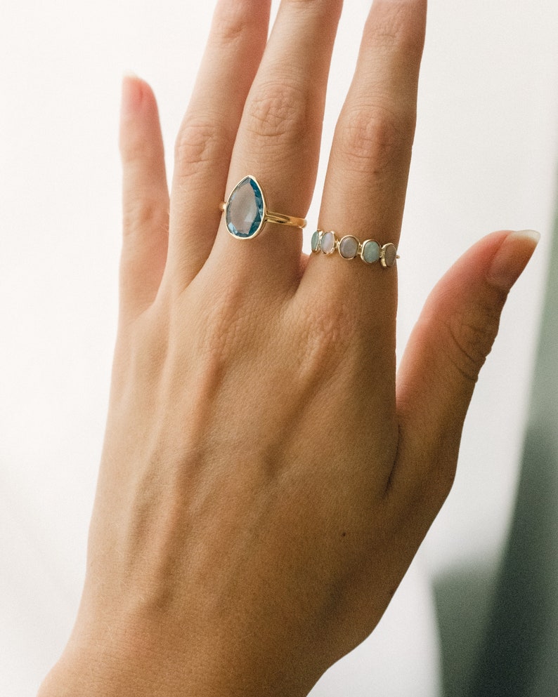 May include: A hand wearing two gold rings. The first ring features a teardrop-shaped blue gemstone. The second ring has a band of small, round, pastel-colored gemstones.