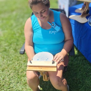 May include: A wooden embroidery hoop stand with a light blue fabric stretched across the hoop. The stand is held by a person wearing a turquoise tank top with the name "Kay" written on it. The background is blurred.