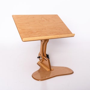 May include: A wooden adjustable stand with a light wood finish. The stand has a flat surface for holding books or other items. The stand is adjustable in height and angle.