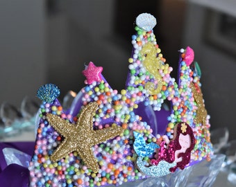 May include: Purple crown decorated with colorful sprinkles, a gold glitter starfish, a mermaid, and other sea-themed embellishments.