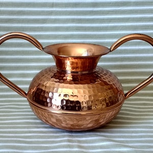 May include: A copper vase with a hammered finish and two arched handles. The vase has a rounded body and a flared top. The handles curve upwards from the sides. The vase is likely for decorative use, perhaps for flowers.