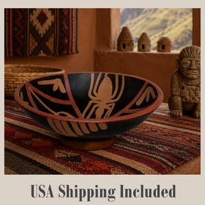 May include: A shallow, round ceramic bowl featuring a matte black interior decorated with a stylized tan spider graphic and red geometric accents. The bowl has a wide, terracotta-colored rim and sits on a small, circular pedestal base.