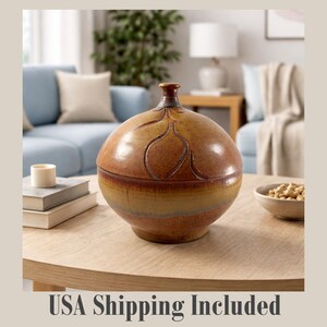 May include: A round, lidded ceramic jar with a brown and yellow glaze. The jar features a decorative leaf-like pattern carved into the surface. It sits on a light-colored wooden table, with a blurred living room background.