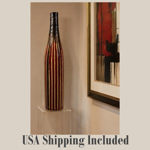 May include: A tall, slender cylindrical vase featuring a dark, textured neck that transitions into a body decorated with vertical stripes in deep red and cream. The vase is positioned upright on a clear, square acrylic pedestal stand.