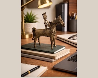 May include: A bronze-colored metal sculpture of a person riding a horse. The sculpture is detailed with intricate patterns and stands on a stack of books. The scene is set on a wooden desk with a laptop, pen, and lamp.