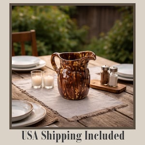 May include: A brown and tan ceramic pitcher with a handle and spout sits on a wooden table. The pitcher is surrounded by plates, candles, and salt and pepper shakers. The table is covered with a linen runner.
