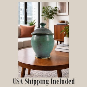 May include: A teal ceramic jar with a lid, sitting on a round wooden table. The jar has a rounded body and a dark gray lid with a small knob. The jar is a decorative item.