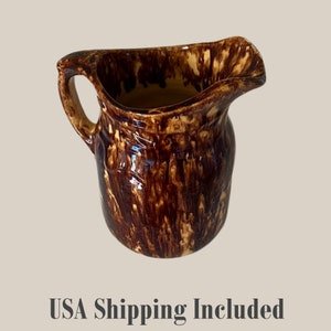 May include: A brown and tan ceramic pitcher with a handle and spout. The pitcher has a glossy finish with a dripped glaze effect. The overall shape is rounded with a slightly flared top.