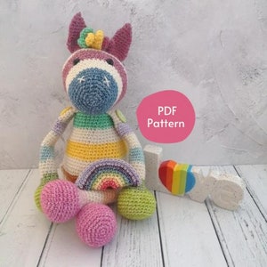 Op de afbeelding: Een kleurrijk gehaakt zebra-speelgoed met een regenboog in zijn hand. Het speelgoed zit op een wit houten oppervlak met een wit bord waarop "LOVE" staat met een regenboog hart.