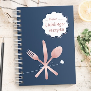 Könnte beinhalten: Ein blaues Spiralblock mit einem weißen Kreis, der den Text "... Meine ... Lieblings-rezepte" in rosa enthält. Das Notizbuch hat ein rosafarbenes Gabel-, Messer- und Löffeldesign mit einem kleinen weißen Herz.