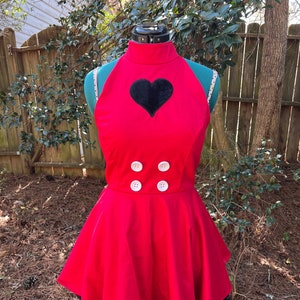 Può includere: Un vestito rosso con un cuore nero sul davanti e tre bottoni bianchi. Il vestito ha un bordo nero intorno al fondo e un collo halter.