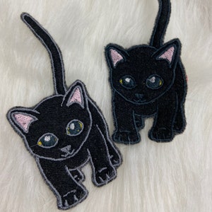 Puede incluir: Dos parches de gato negro con detalles bordados. Los gatos tienen ojos grandes y expresivos, orejas internas rosas y contornos grises. Los parches están sobre un fondo blanco y esponjoso.