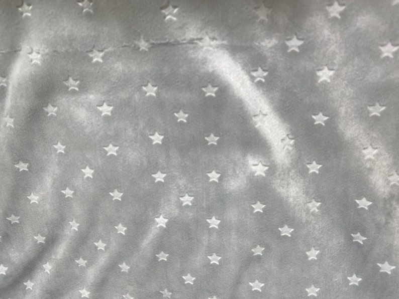 May include: A close-up of a soft, gray fabric with a pattern of white stars. The stars are evenly spaced across the surface of the fabric. The fabric has a velvety texture and a subtle sheen.
