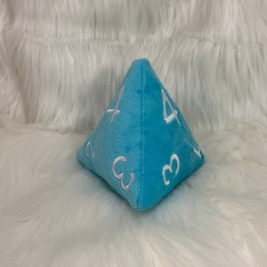 May include: A light blue plush dice shaped like a pyramid with white embroidered numbers on each side. The dice is sitting on a white fluffy surface.