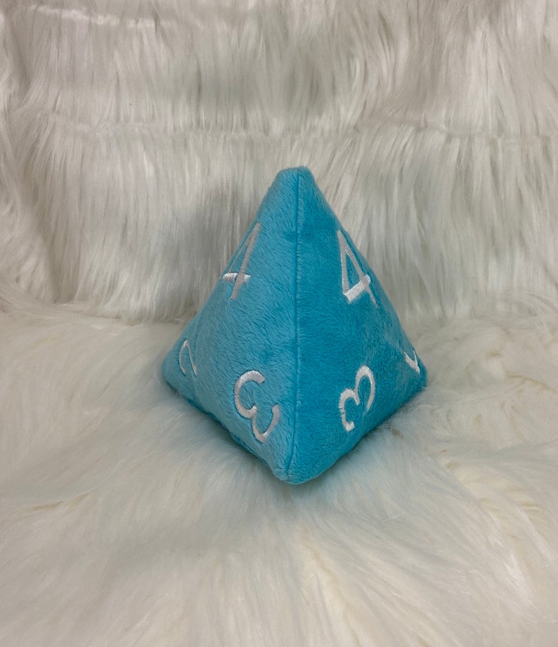 May include: A light blue plush dice shaped like a pyramid with white embroidered numbers on each side. The dice is sitting on a white fluffy surface.
