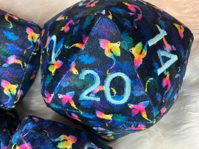 May include: A plush, black, 20-sided dice with a colorful, iridescent pattern of dragons. The numbers are embroidered in white.