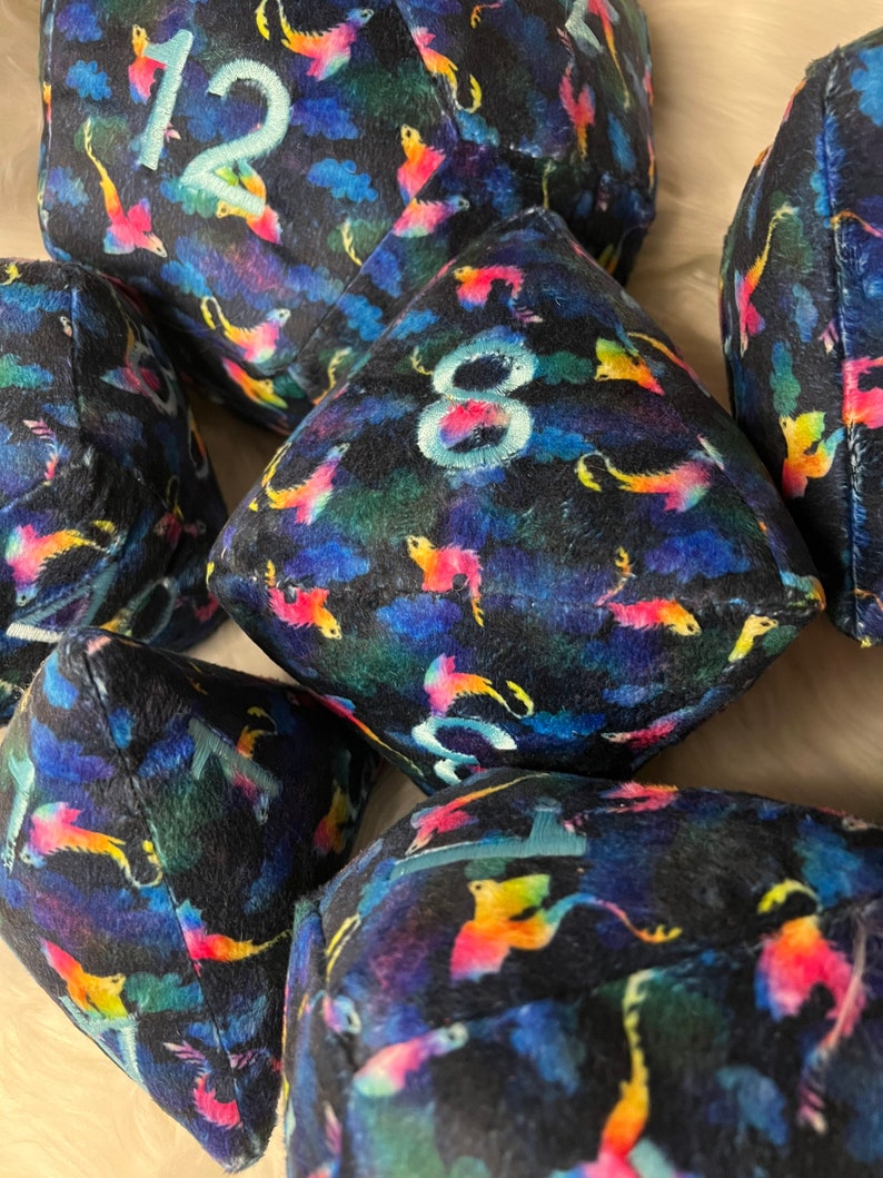 May include: A set of six plush dice with a dark blue background and a colorful, abstract pattern of birds. The dice are numbered 1, 2, 3, 4, 5, and 6.