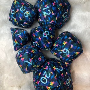May include: A set of seven plush dice with a dark blue background and a colorful, abstract pattern. The dice are numbered 1, 2, 4, 6, 8, 10, and 20.