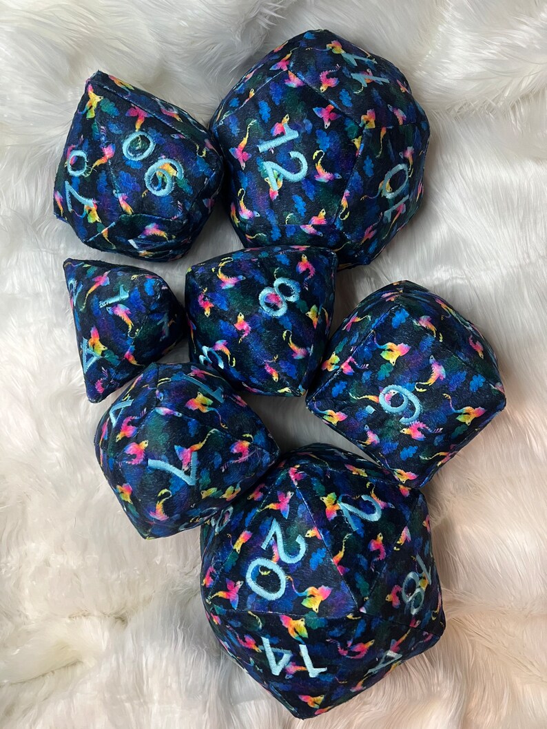 May include: A set of seven plush dice with a dark blue background and a colorful, abstract pattern. The dice are numbered 1, 2, 4, 6, 8, 10, and 20.