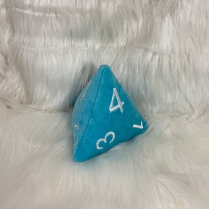 May include: A light blue plush dice with white numbers 1, 3, 4, and 5. The dice is a D4, or four-sided die.