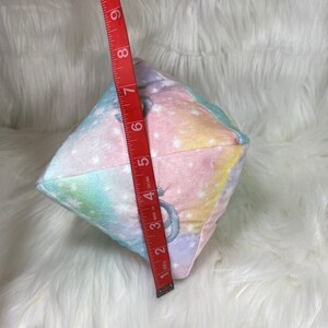 May include: A plush, multi-colored cube with a celestial pattern of pastel colors and white stars. The cube is approximately 6 inches in height. The cube has a silver embroidered design on each side.