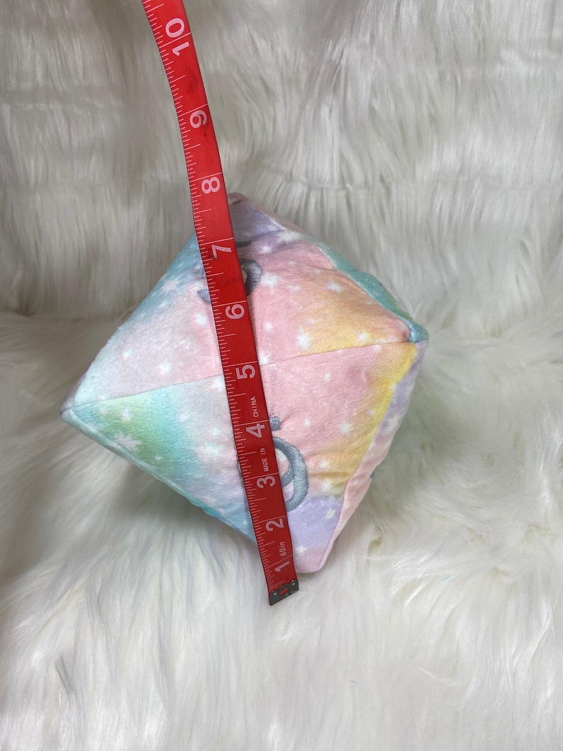 May include: A plush, multi-colored cube with a celestial pattern of pastel colors and white stars. The cube is approximately 6 inches in height. The cube has a silver embroidered design on each side.