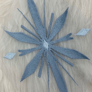 Puede incluir: Un diseño de copo de nieve azul claro bordado sobre un fondo de tela blanca.